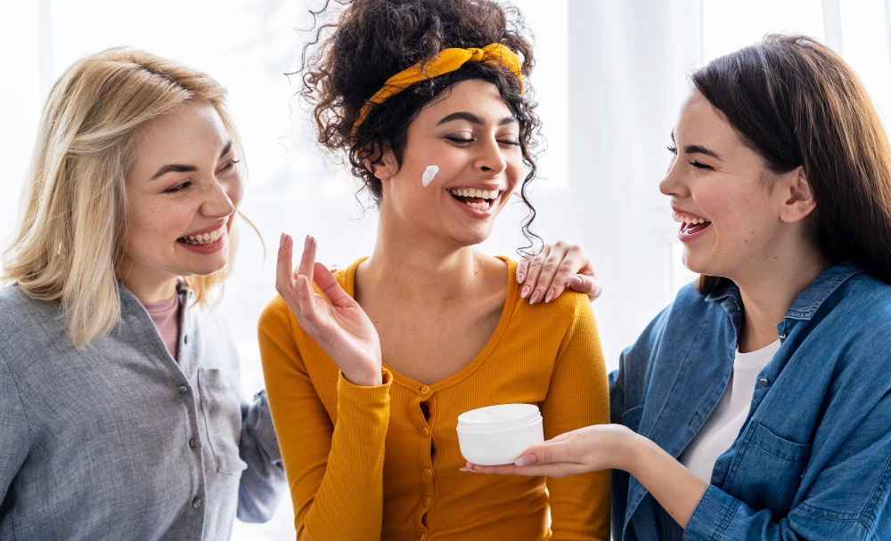 Three friends laughing and sharing skincare tips while applying cream.